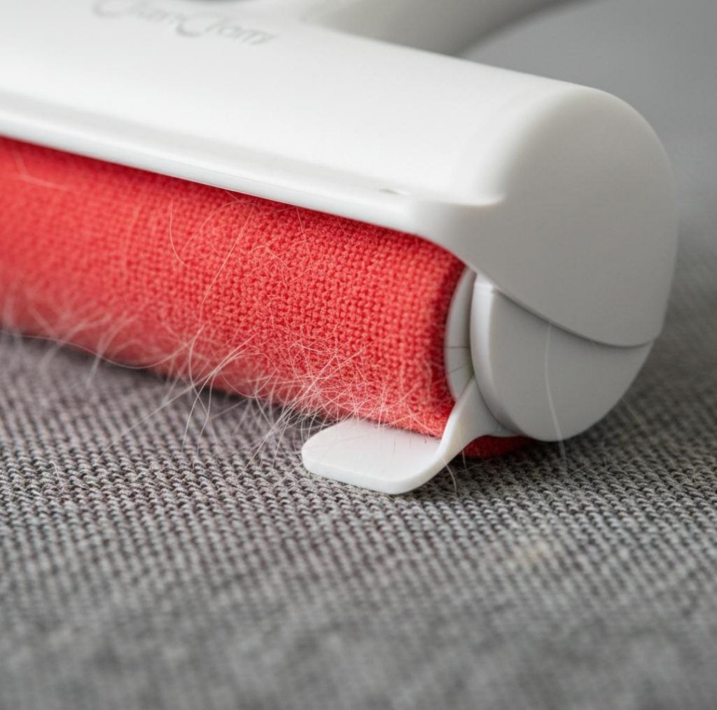 A macro close-up shot of the ChomChom Roller's red microfiber surface and white squeegee mechanism trapping white pet hair from a gray textured fabric sofa.